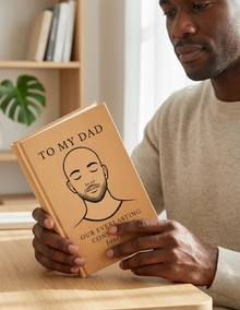 Man holding a book titled 'To My Dad: Our Everlasting Connection Journal' with a personal illustration on a wooden surface.