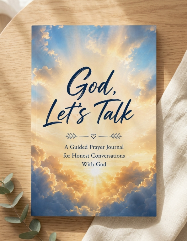 A guided prayer journal titled 'God, Let's Talk' lies flat on a light wood surface with a soft white cloth and eucalyptus leaves around it. The cover features a dramatic sky with blue and gold clouds and dark navy text.