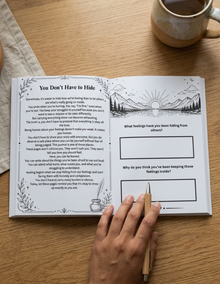 An open journal or notebook spread on a wooden table, showing a poem titled 'You Don't Have to Hide' on the left page and reflective prompts on the right. A hand holds a wooden pencil over the pages, with a ceramic mug partially visible in the upper right.